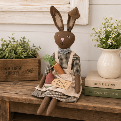 A rustic fabric bunny doll sits on a wooden bench in a styled farmhouse setting. The doll has brown ears and a brown face, wearing a black-and-white gingham dress and apron over a gray ruffled collar, and holds a carrot and a pinkish root vegetable.