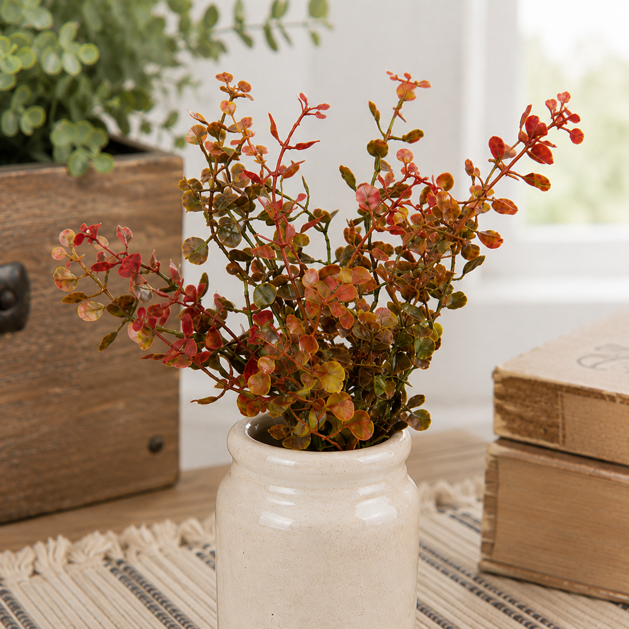 Artificial autumn foliage with red, orange, and green leaves in a small white ceramic jar, styled on a table with a woven runner, a wooden box, and books in a bright room.