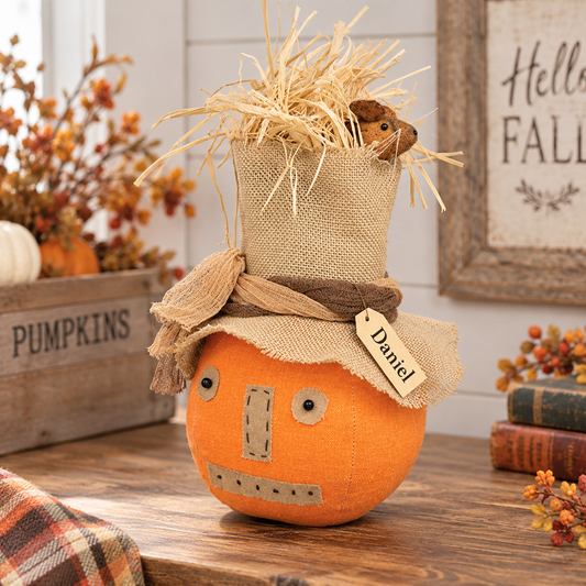 An orange fabric pumpkin decoration with a handmade straw hat and burlap scarf, displayed on a wooden table in a fall-themed setting with a 'Hello Fall' sign and a wooden crate labeled 'PUMPKINS'.