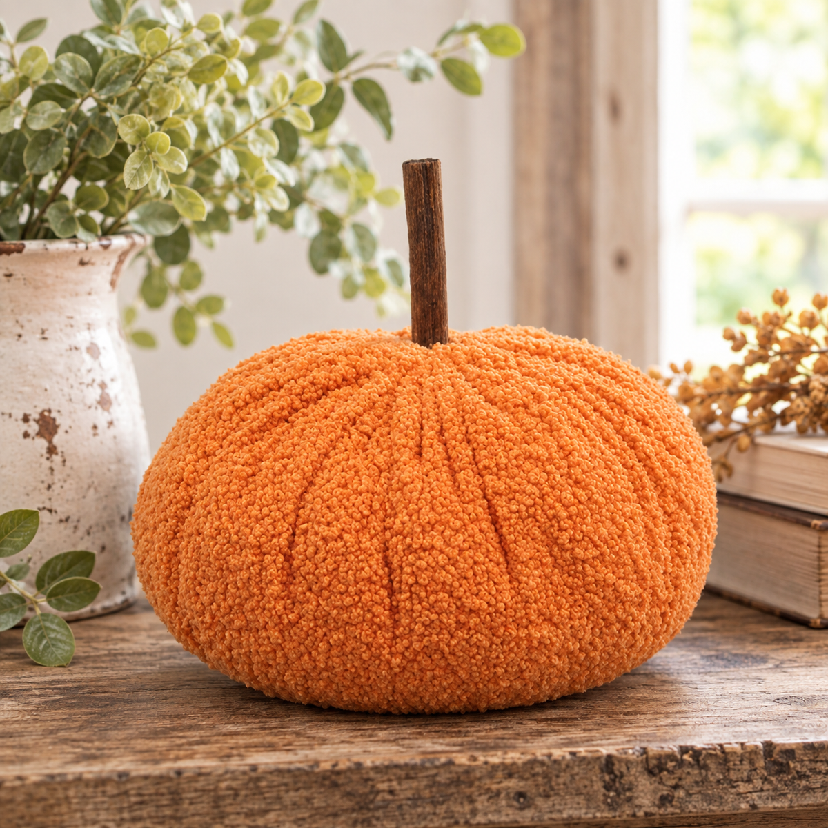 An orange textured fabric pumpkin decoration with a brown stem, displayed on a rustic wooden table with a white ceramic vase of greenery and books in the background.