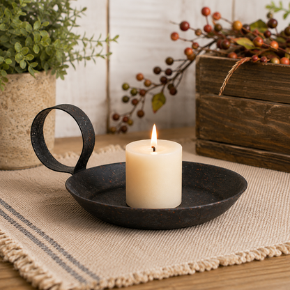 A black metal candle holder with a loop handle and a lit white pillar candle, styled on a woven placemat with rustic decorative elements in the background.