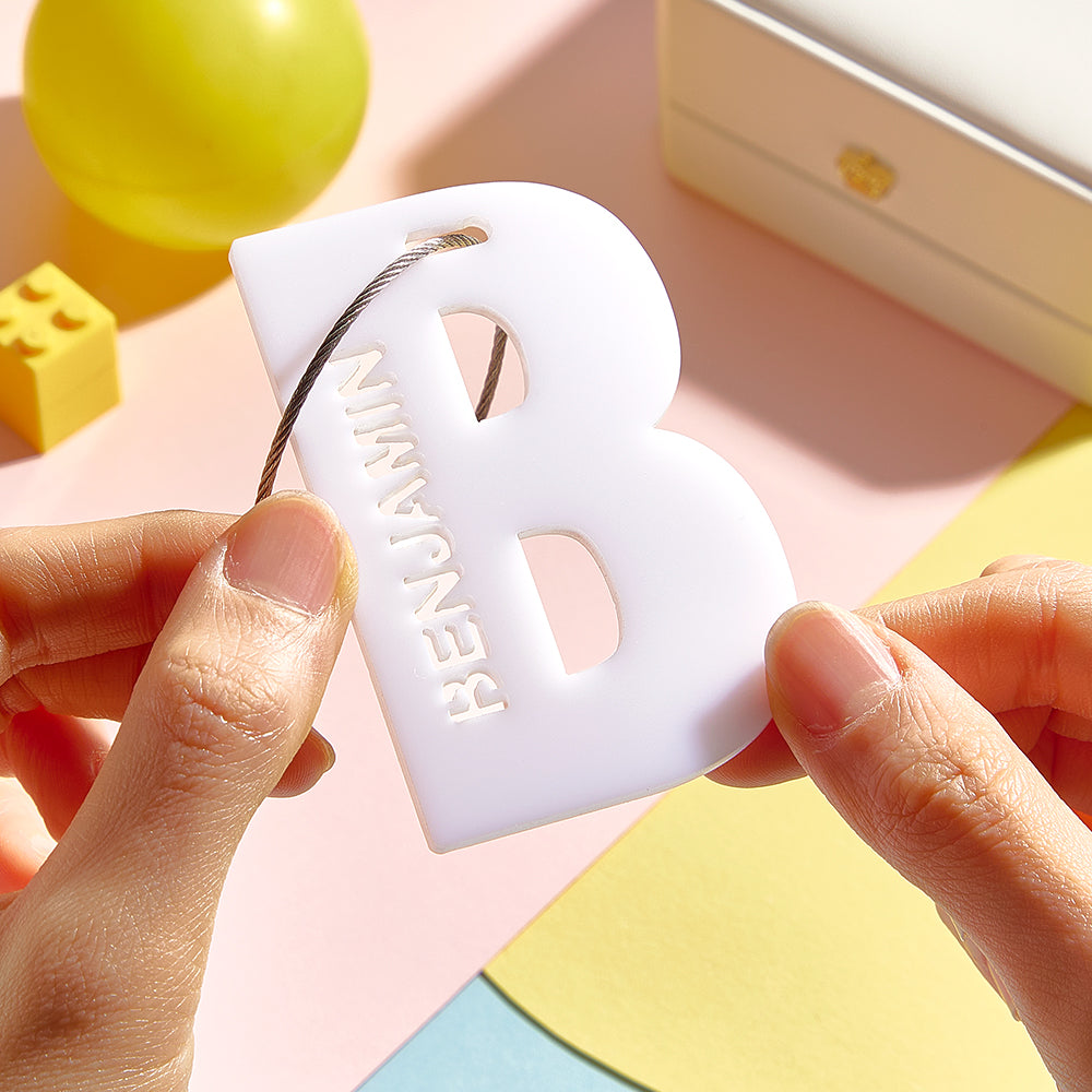 A white 3D printed luggage tag in the shape of the letter "B", showcasing its sleek design and customizable feature for personalized use on school bags or luggage.