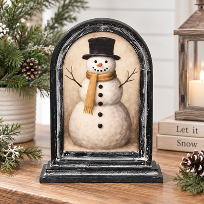 A decorative tabletop snowman figurine in an arched black frame, styled on a wooden surface with holiday greenery, pinecones, a lit candle, and stacked books in the background.