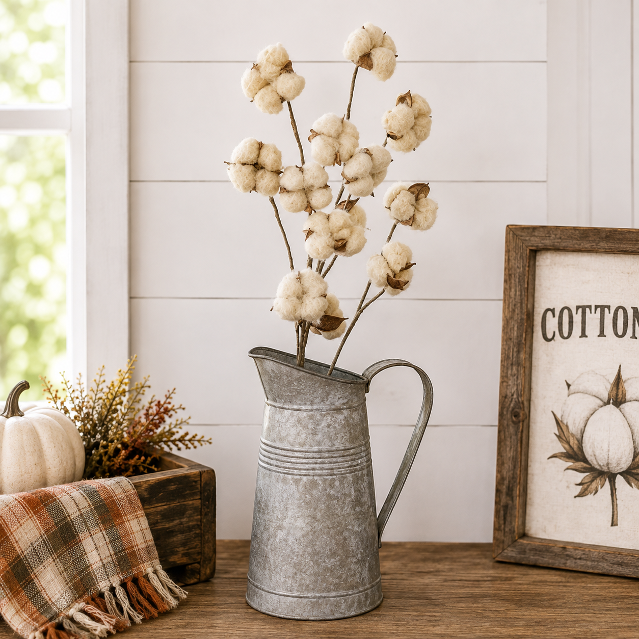 A galvanized metal pitcher vase filled with dried cotton stems, styled on a wooden table next to a framed cotton print and a small pumpkin in a rustic home decor setting.
