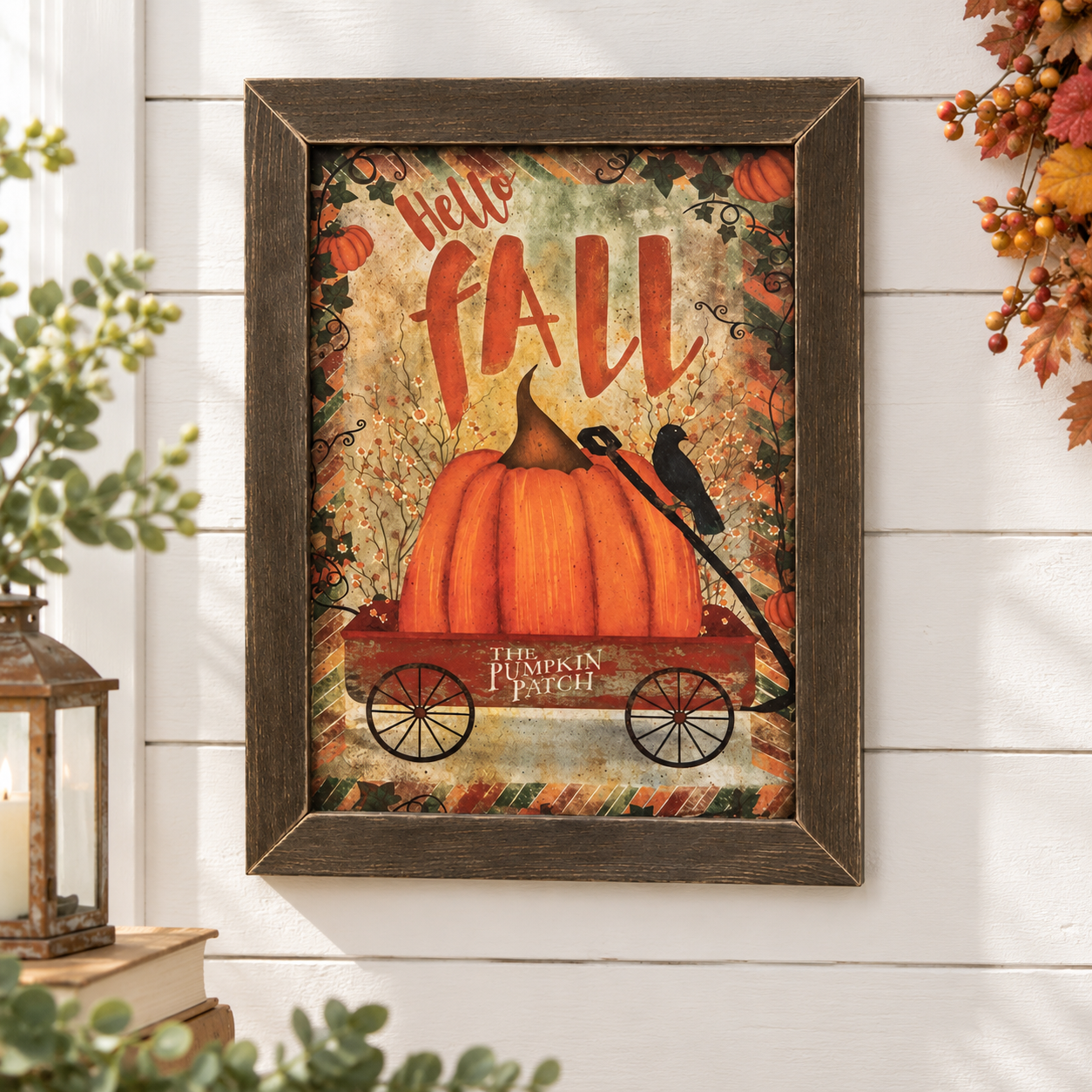 Framed autumn wall art hanging on a white shiplap wall, featuring a large orange pumpkin in a red wagon with the words “Hello FALL” and “The Pumpkin Patch,” surrounded by fall foliage and a crow. Decorative plants and a lantern are visible at the sides.
