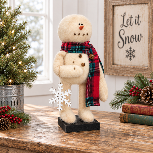 A plush cream-colored snowman figurine wearing a red, green, and black plaid scarf, standing on a black base on a wooden table. The scene is decorated for Christmas with a small lit tree, books, pinecones, and a framed sign reading 'Let it Snow'.