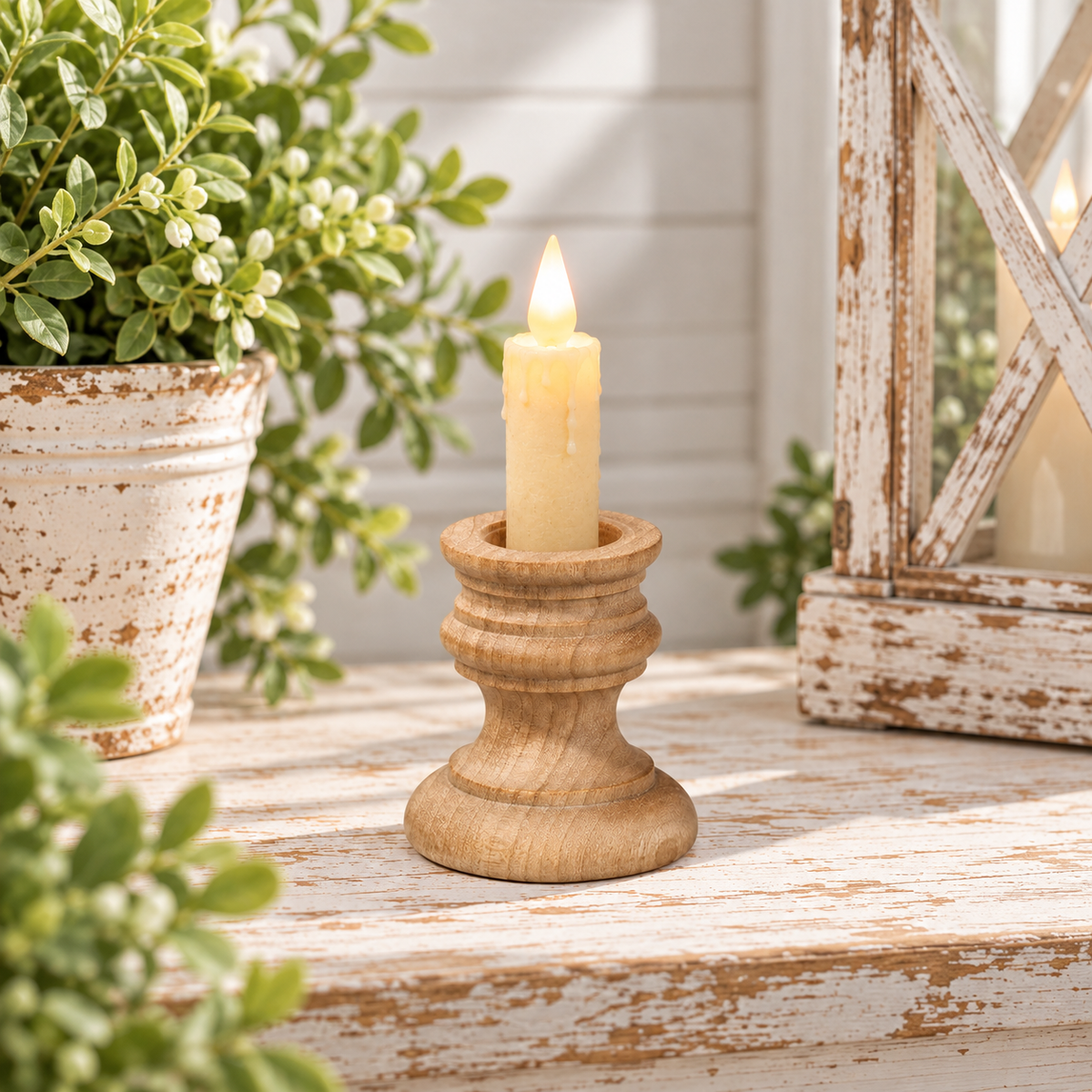A small natural wood candle holder with a lit cream-colored pillar candle, styled on a distressed white wooden surface with potted green plants and a lantern in the background.