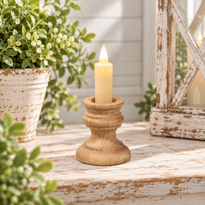 A small natural wood candle holder with a lit cream-colored pillar candle, styled on a distressed white wooden surface with potted green plants and a lantern in the background.