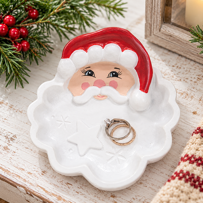 A white ceramic Santa Claus head ring holder with a red hat, shown on a rustic white wooden surface with holiday greenery and rings placed in the center.