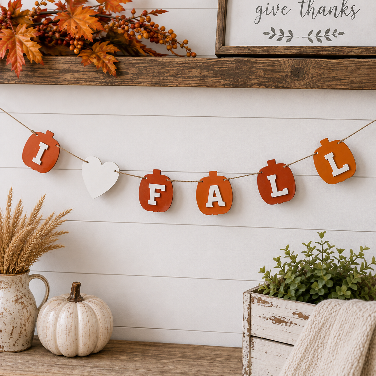 A rustic autumn-themed 'I FALL' banner garland hanging on a white shiplap wall above a wooden shelf. The banner features orange pumpkin-shaped letters with white letters inside, a white heart, and other orange autumn decorative elements.