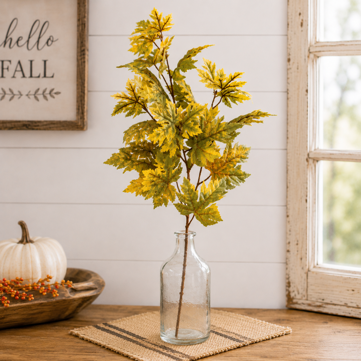 Artificial autumn maple leaves in a clear glass bottle vase, styled on a woven placemat on a wooden table with a white pumpkin and a rustic framed sign in the background.