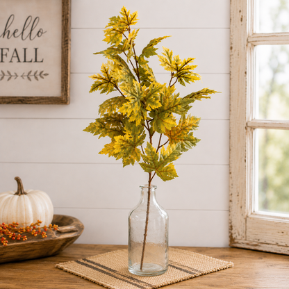 Artificial autumn maple leaves in a clear glass bottle vase, styled on a woven placemat on a wooden table with a white pumpkin and a rustic framed sign in the background.
