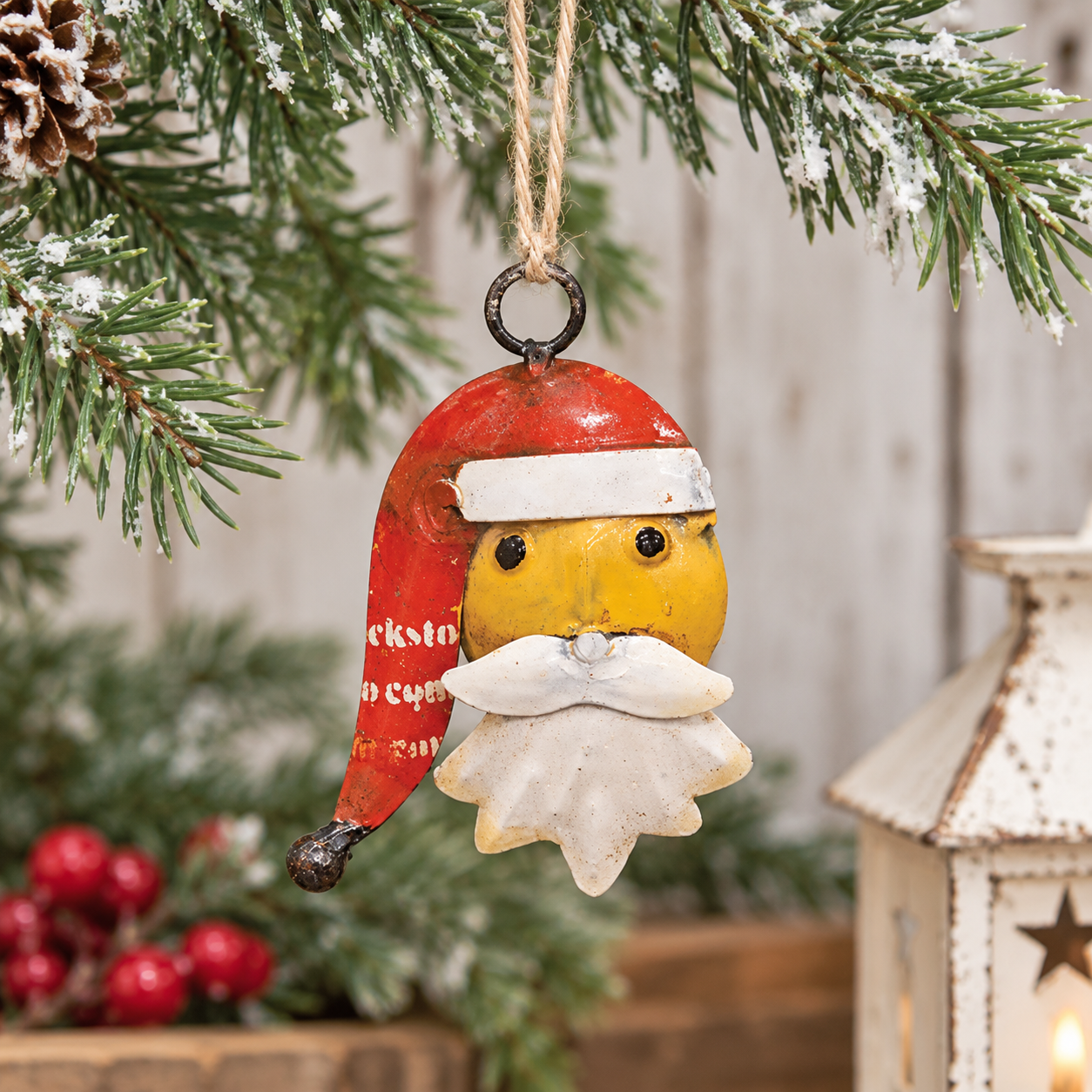 A rustic Christmas ornament shaped like a stylized Santa face hanging from a pine branch. The ornament has a red hat with white trim, a yellow face and beard, and is shown against a softly blurred holiday background with a lantern and red berries.