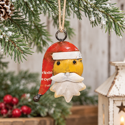 A rustic Christmas ornament shaped like a stylized Santa face hanging from a pine branch. The ornament has a red hat with white trim, a yellow face and beard, and is shown against a softly blurred holiday background with a lantern and red berries.