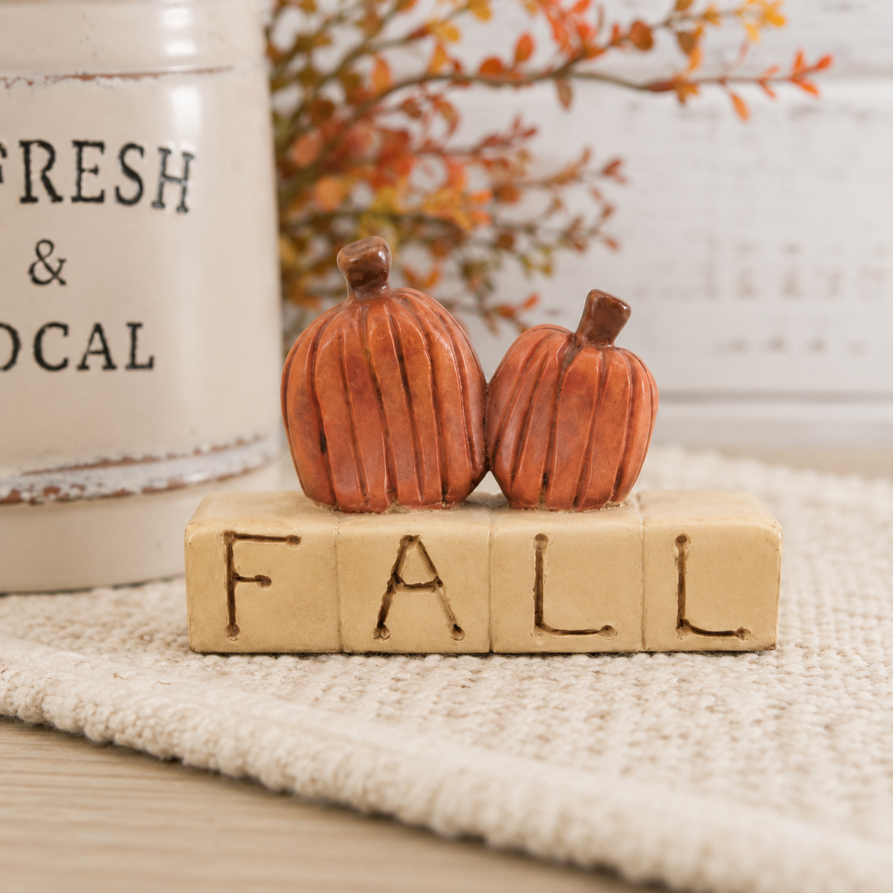 A decorative fall-themed tabletop piece with two small orange-brown pumpkin figurines standing on a beige block spelling FALL, styled on a textured cream cloth with a blurred white canister and autumn branches in the background.