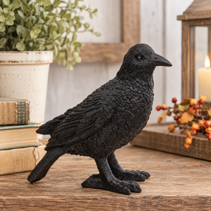 A black crow figurine with highly detailed textured feathers, standing on a rustic wooden surface with a potted plant and books in the background.