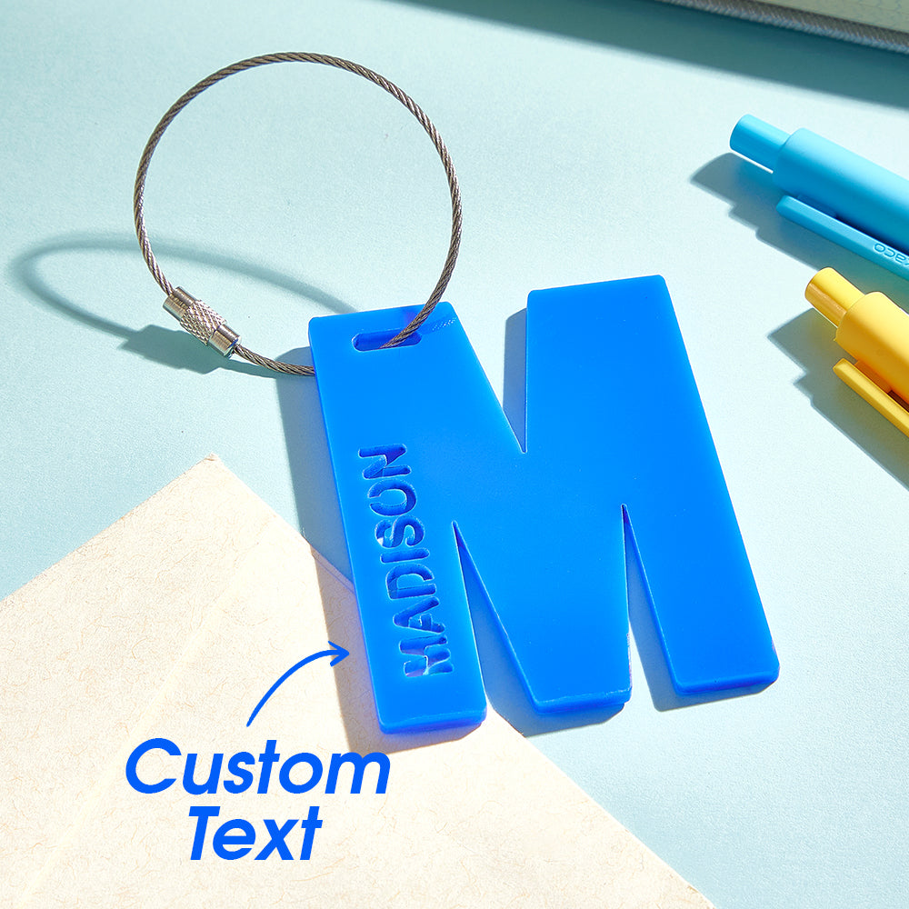 A close-up view of a 3D printed luggage tag shaped like the letter "M" in blue, with a text overlay that says "Custom text," emphasizing its vibrant color and personalized design.