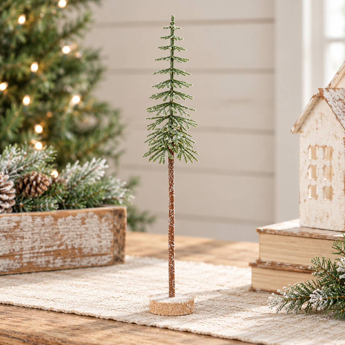A small decorative artificial Christmas tree with a brown textured trunk and green needle-like branches, standing on a round base on a rustic table. The background shows a softly blurred holiday scene with a lit Christmas tree, pinecones, books, and a small house ornament.