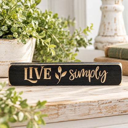 A rectangular black wooden sign with the words 'LIVE simply' in a light natural wood tone, styled on a rustic white wooden surface with green plants and books in the background.
