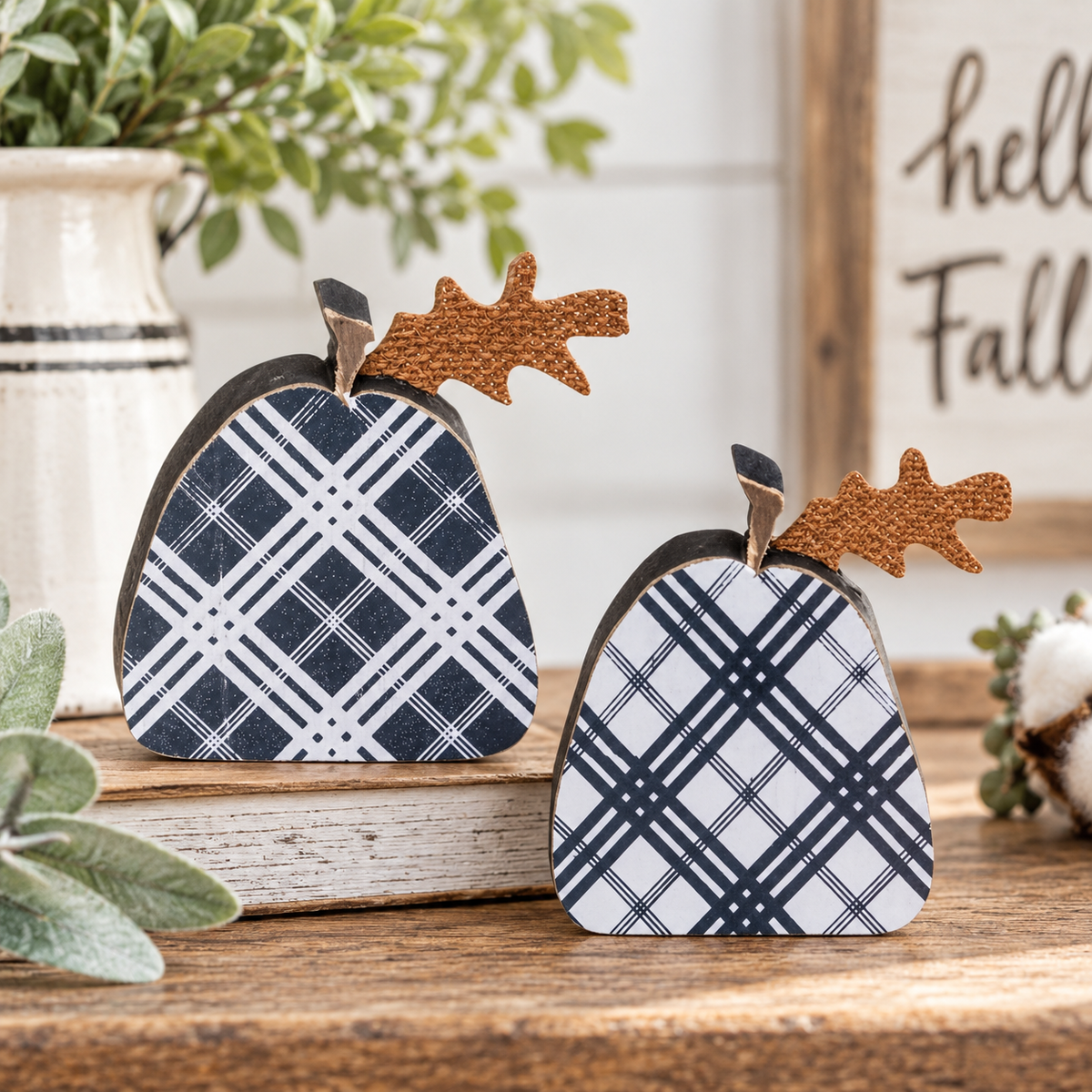 Two decorative fall pumpkins with navy and white plaid patterns and brown faux-wood stems, styled on a rustic wooden table with a book, potted plant, and a sign in the background.