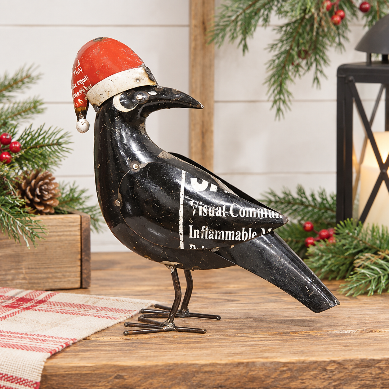 A black metal raven figurine wearing a red Santa hat, displayed on a wooden surface with holiday greenery and a lantern in the background.