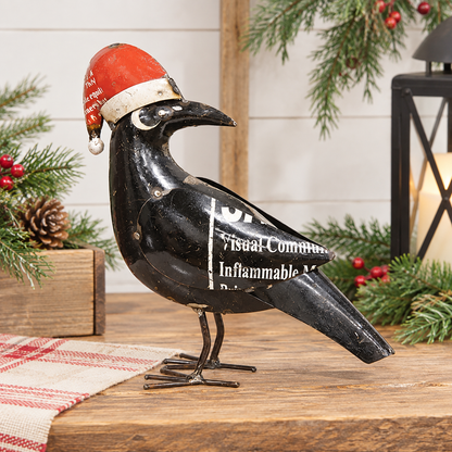 A black metal raven figurine wearing a red Santa hat, displayed on a wooden surface with holiday greenery and a lantern in the background.