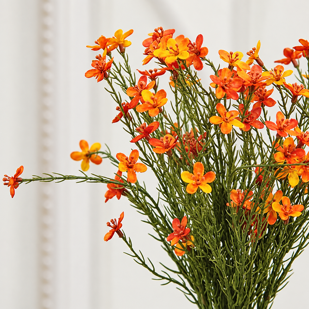 A bouquet of artificial orange and yellow flowers with green stems, arranged against a soft white background.