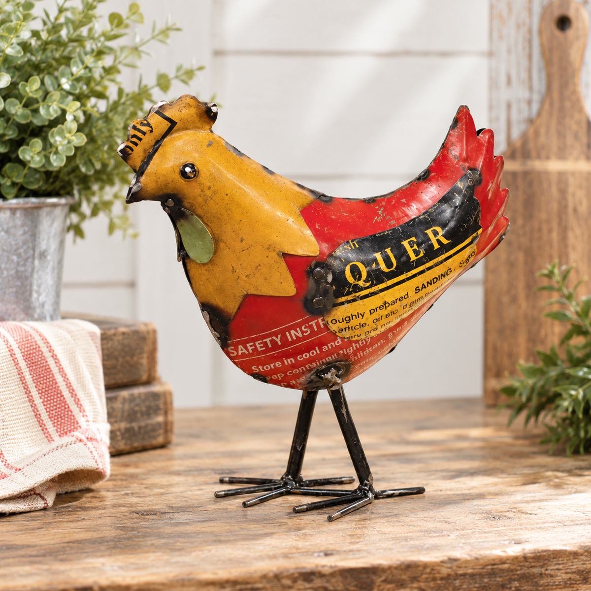 A vintage-style metal rooster figurine with a distressed yellow, red, and black paint scheme, standing on a rustic wooden surface with a kitchen-themed background.