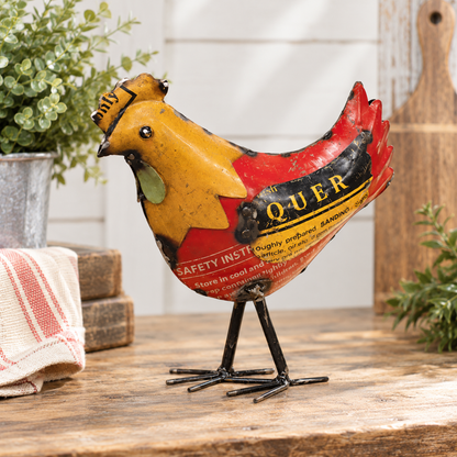 A vintage-style metal rooster figurine with a distressed yellow, red, and black paint scheme, standing on a rustic wooden surface with a kitchen-themed background.