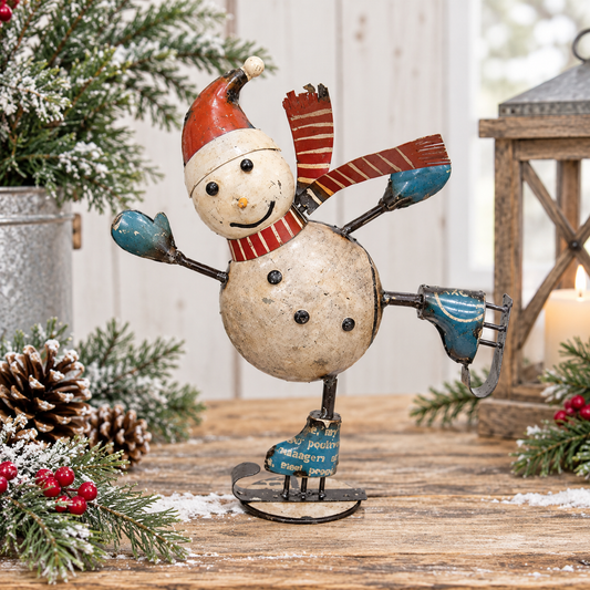 A rustic metal snowman figurine posed on a small ice skate, decorated with a red and white striped hat and scarf, blue hands and boots, and black buttons, displayed on a wooden surface with holiday greenery and a lantern in the background.
