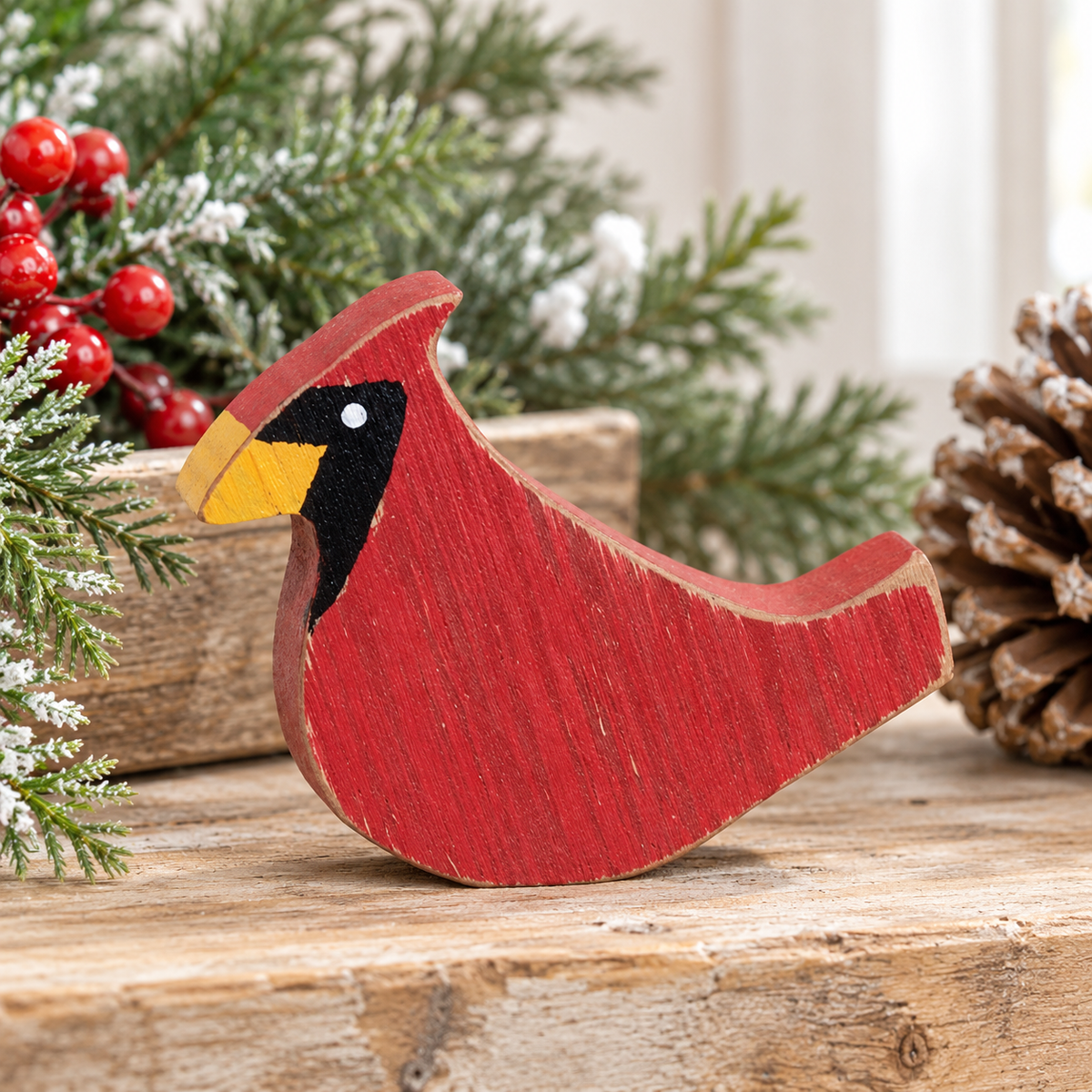 A red wooden cardinal bird figurine with a yellow beak, black head, and white eye, displayed on a rustic wooden surface with holiday greenery, red berries, and a pinecone in the background.