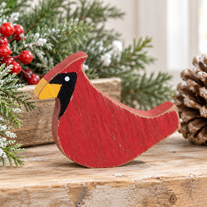 A red wooden cardinal bird figurine with a yellow beak, black head, and white eye, displayed on a rustic wooden surface with holiday greenery, red berries, and a pinecone in the background.