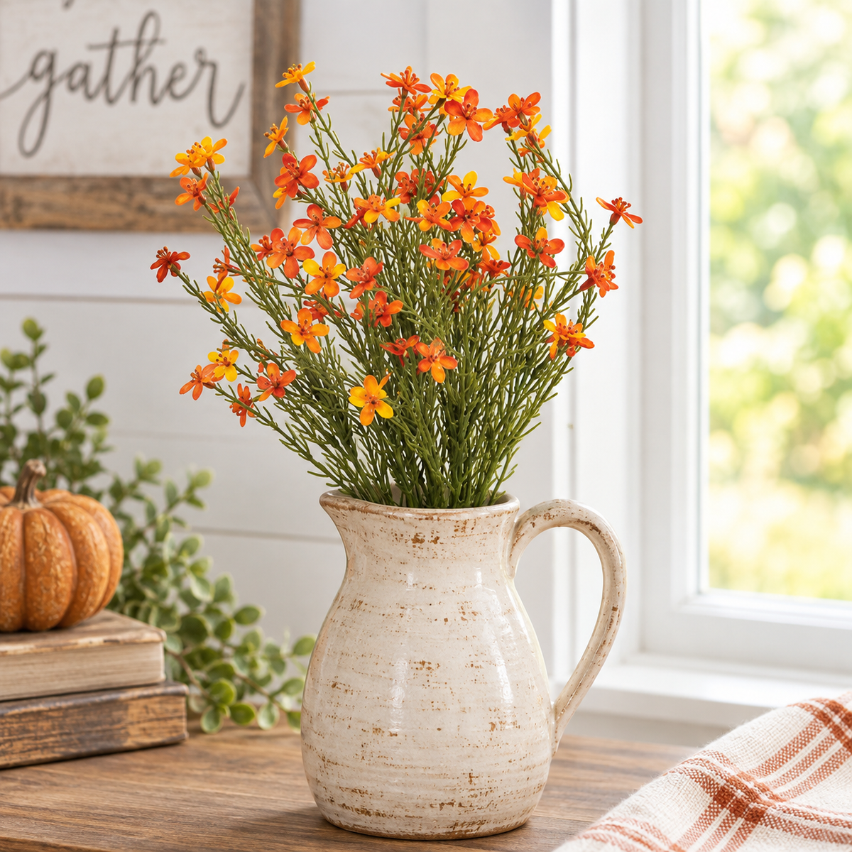 A rustic ceramic pitcher vase filled with orange and yellow faux flowers, styled on a wooden table next to a small pumpkin and greenery in front of a window.