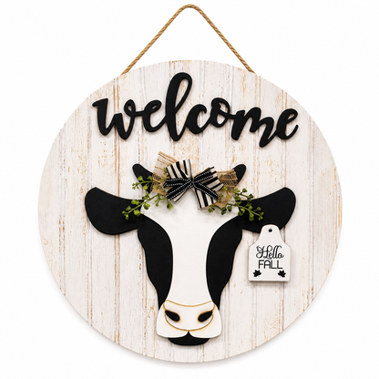 Round wooden welcome sign with black and white cow, raised welcome lettering, jute hanger, and interchangeable seasonal ear tag for farmhouse wall or door décor