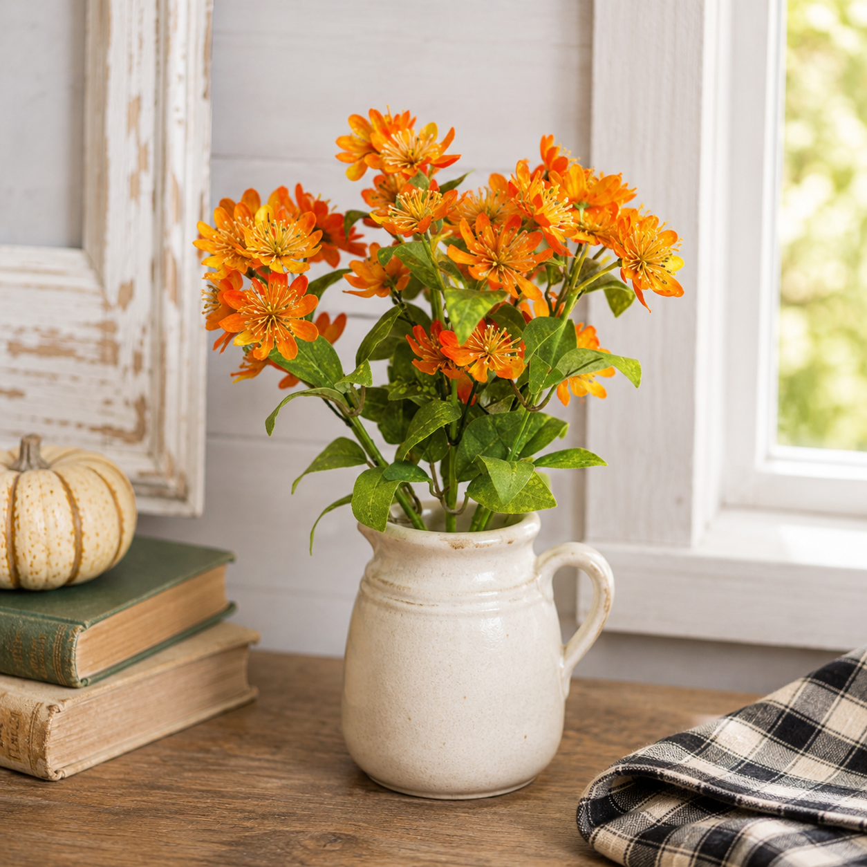 A bouquet of bright orange marigold-like flowers with green leaves arranged in a small cream ceramic pitcher, placed on a wooden table in a rustic room setting with books, a small pumpkin, and a window in the background.