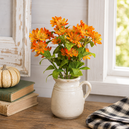 A bouquet of bright orange marigold-like flowers with green leaves arranged in a small cream ceramic pitcher, placed on a wooden table in a rustic room setting with books, a small pumpkin, and a window in the background.