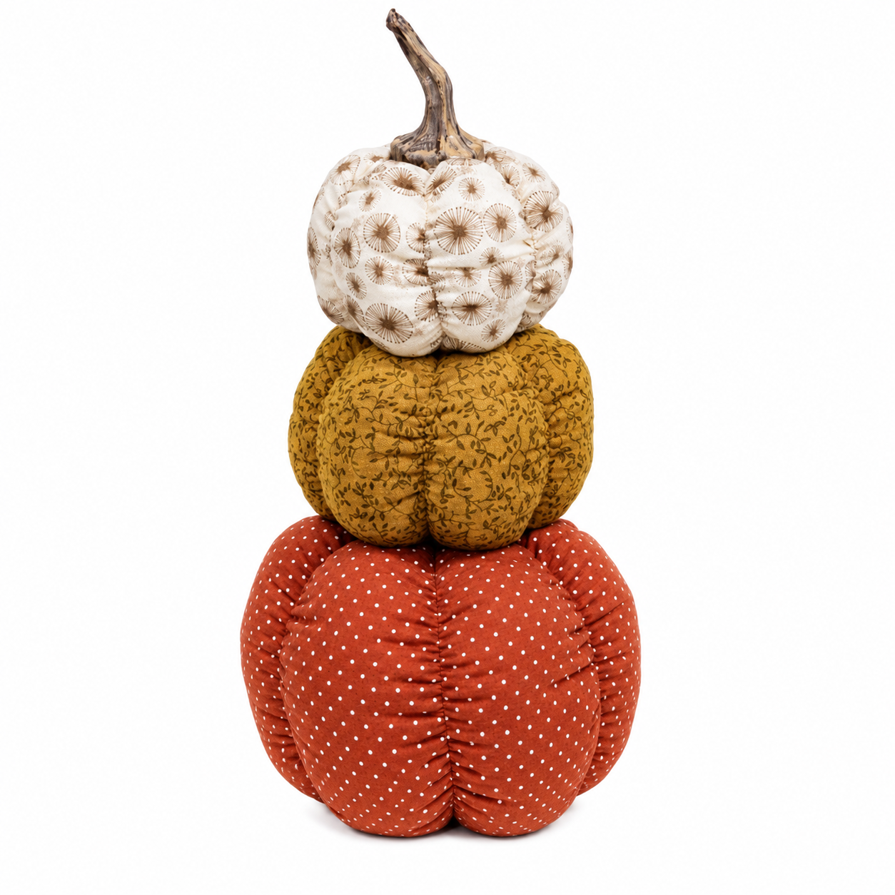 Three stacked fabric decorative pumpkins on a plain white background. The top pumpkin is off-white with brown spots and a brown stem, the middle pumpkin is mustard yellow with a dark leafy pattern, and the bottom pumpkin is rust orange with small white polka dots.