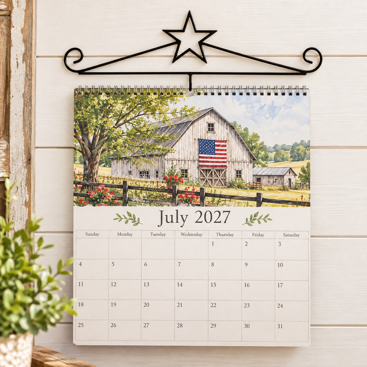 A spiral-bound July 2027 desk calendar hanging from a black metal wall hook. The calendar cover features a watercolor painting of a white barn with an American flag, trees, and a fence, set against a white wood-paneled wall with a small green plant in the foreground.