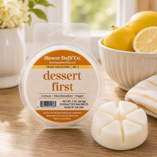 A round plastic tub of Slower Days Co. wax melts labeled 'dessert first' sits on a wooden surface with a white flower vase on the left and a bowl of lemons on the right.