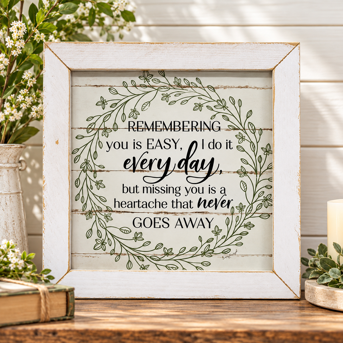 A square framed decorative sign with a rustic white distressed frame and a printed quote surrounded by a green leaf wreath. The sign sits on a wooden surface with flowers, a candle, and books in a styled home decor setting.