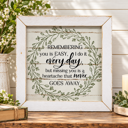 A square framed decorative sign with a rustic white distressed frame and a printed quote surrounded by a green leaf wreath. The sign sits on a wooden surface with flowers, a candle, and books in a styled home decor setting.