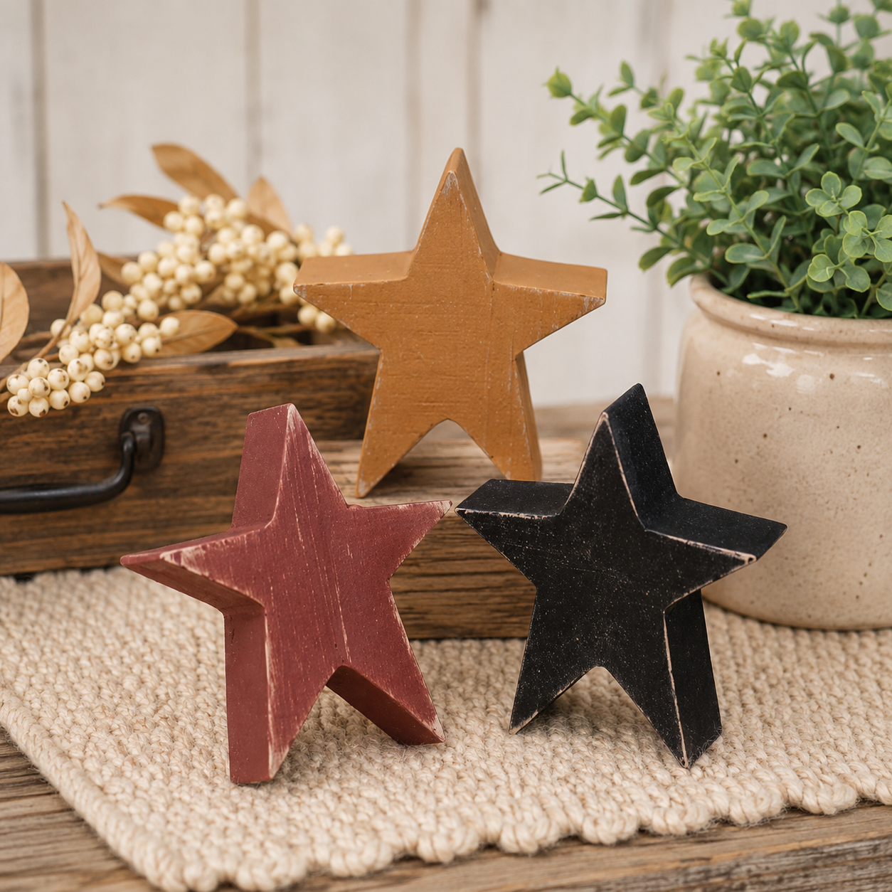 Three rustic wooden star-shaped ornaments in tan, red, and black, displayed on a woven beige runner with a potted plant and decorative box in the background.