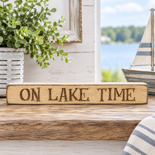 A rectangular wooden sign engraved with 'ON LAKE TIME' in dark brown lettering, displayed on a rustic wooden surface with a blurred background showing a lake, trees, a potted plant, and a small sailboat model.