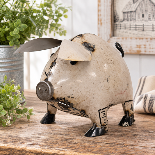 A rustic decorative pig figurine made from recycled metal parts, featuring a distressed off-white body, black and silver accents, and a propeller blade on its head, displayed on a wooden tabletop with a blurred background.