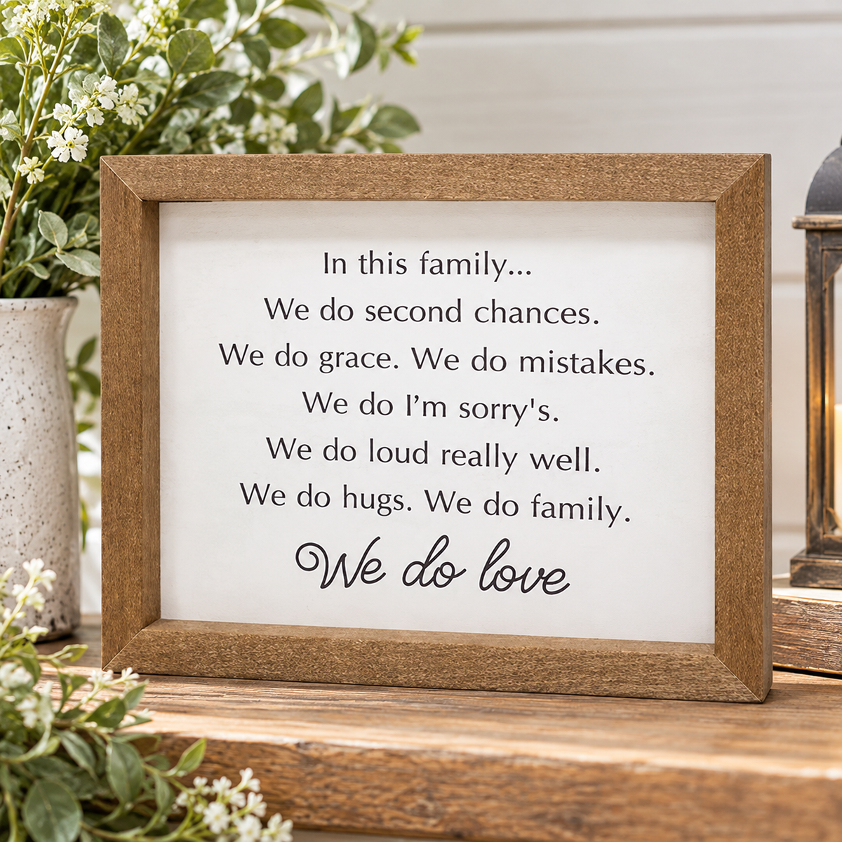 A rustic wooden framed sign with a white background and black text reading a family message about second chances, grace, mistakes, apologies, and love, displayed on a wooden surface with flowers and home decor props.