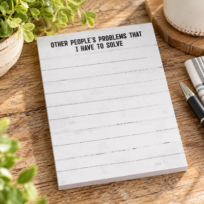 A white notepad with a rustic wood-plank print and the text 'OTHER PEOPLE'S PROBLEMS THAT I HAVE TO SOLVE' in black, placed on a wooden tabletop with plants, a mug, and a pen nearby.