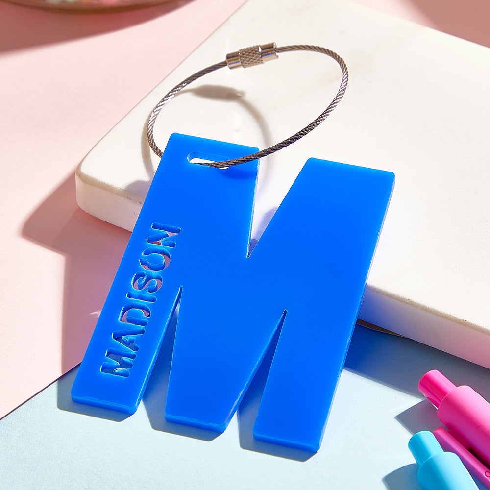 A close-up view of a 3D printed luggage tag shaped like the letter "M" in blue, highlighting its smooth surface and durable acrylic material, perfect for personalized identification.