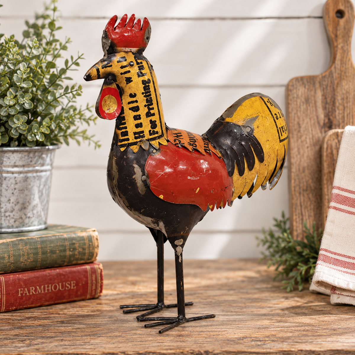 A decorative metal rooster figurine with a distressed finish, featuring red, yellow, black, and brown sections with printed text, displayed on a rustic wooden surface with a potted plant and books in the background.