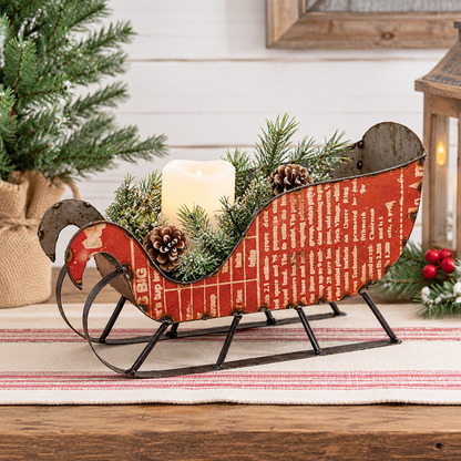 A rustic red metal sleigh-shaped candle holder with black sled runners, filled with green pine branches, pinecones, and a white pillar candle, styled on a striped rug with holiday decor in the background.