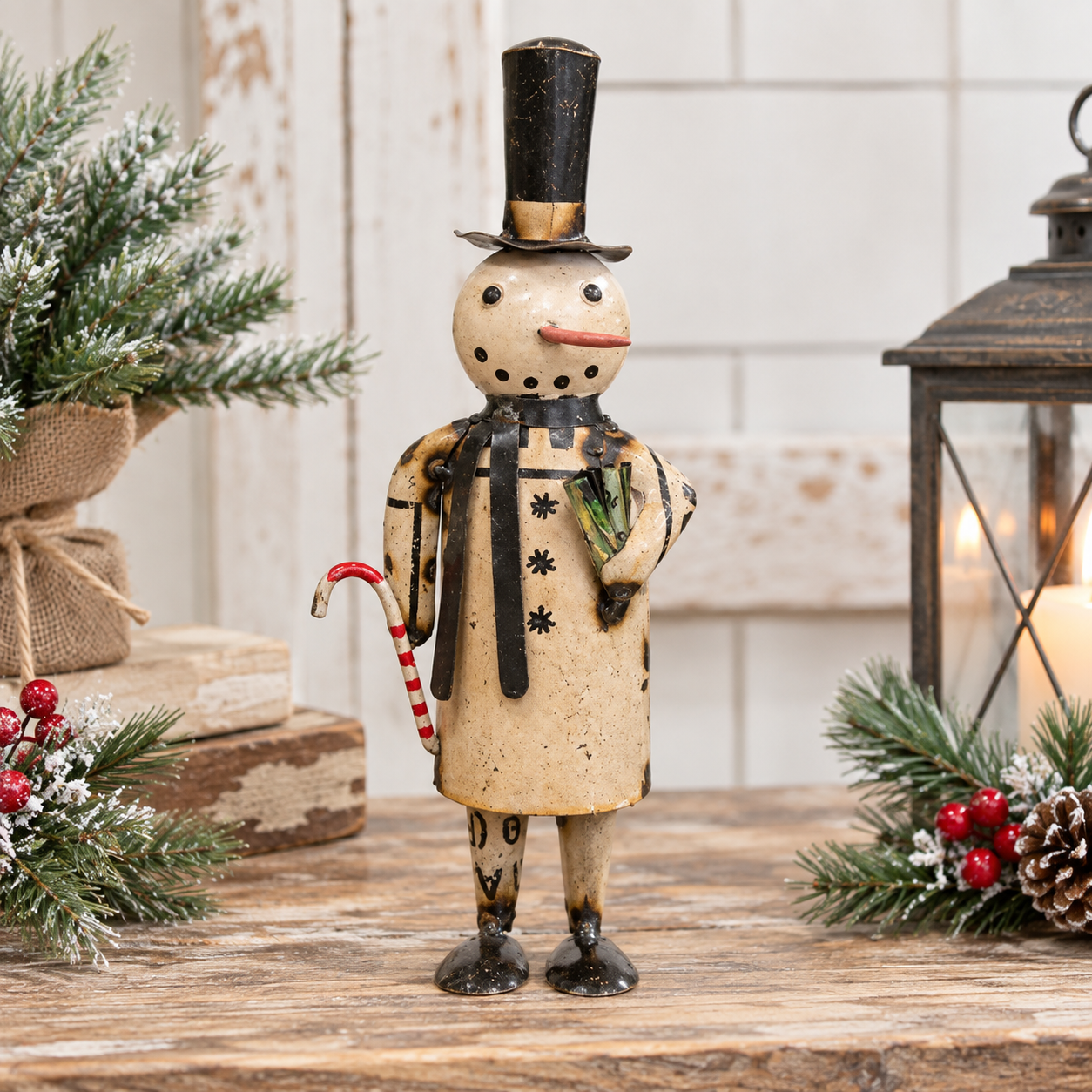 A decorative snowman figurine stands on a rustic wooden surface, wearing a tall black top hat, black-and-beige coat, and holding a red-and-white candy cane. The scene is styled with evergreen branches, red berries, stacked books, and a lantern in the background.